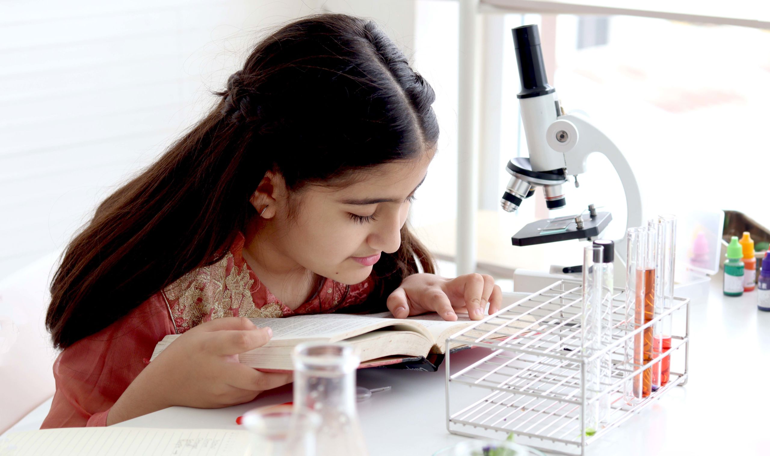 Jente sitter foran et mikroskop og leser i en bok.