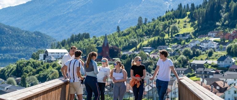 Syv studenter står på et utkikkspunkt på Campus Sogndal, hvor utsikten er store fjell, frodig grønt landskap, og noen hus og en kirke.