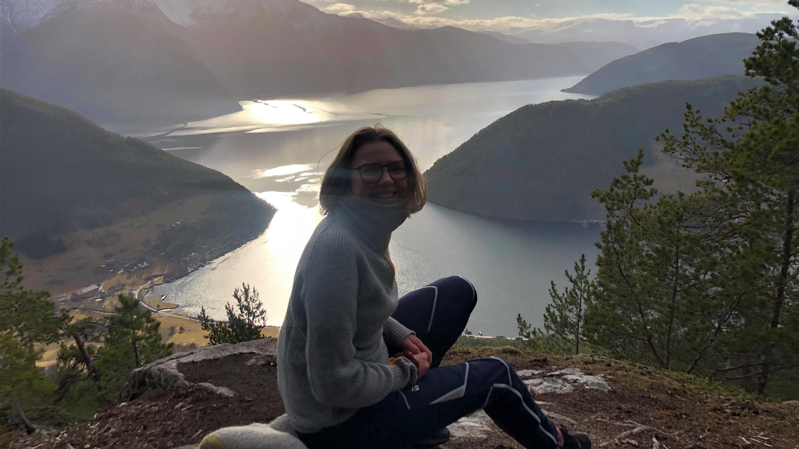 MAri sittende på fjelltopp med utsikt over fjorden. 