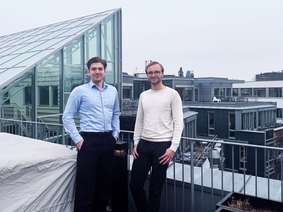 Philip Hodne, ein mørkhåra kar i blå skjorte og svart bukse, står til venstre i bildet. Til høgre står Thomas Solberg Irgens, ein fyr med brunt hår, trimma skjegg og briller, kledd i ein kvit genser og svart bukse. I bakgrunnen ser ein nokre bygningar og ein overskya himmel.