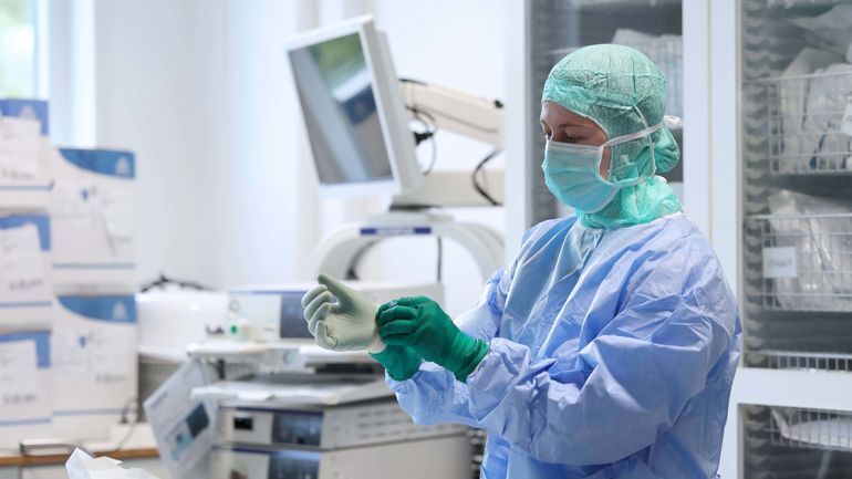 Sykepleier i full uniform, munnbind og hårnett tar på seg plasthansker.