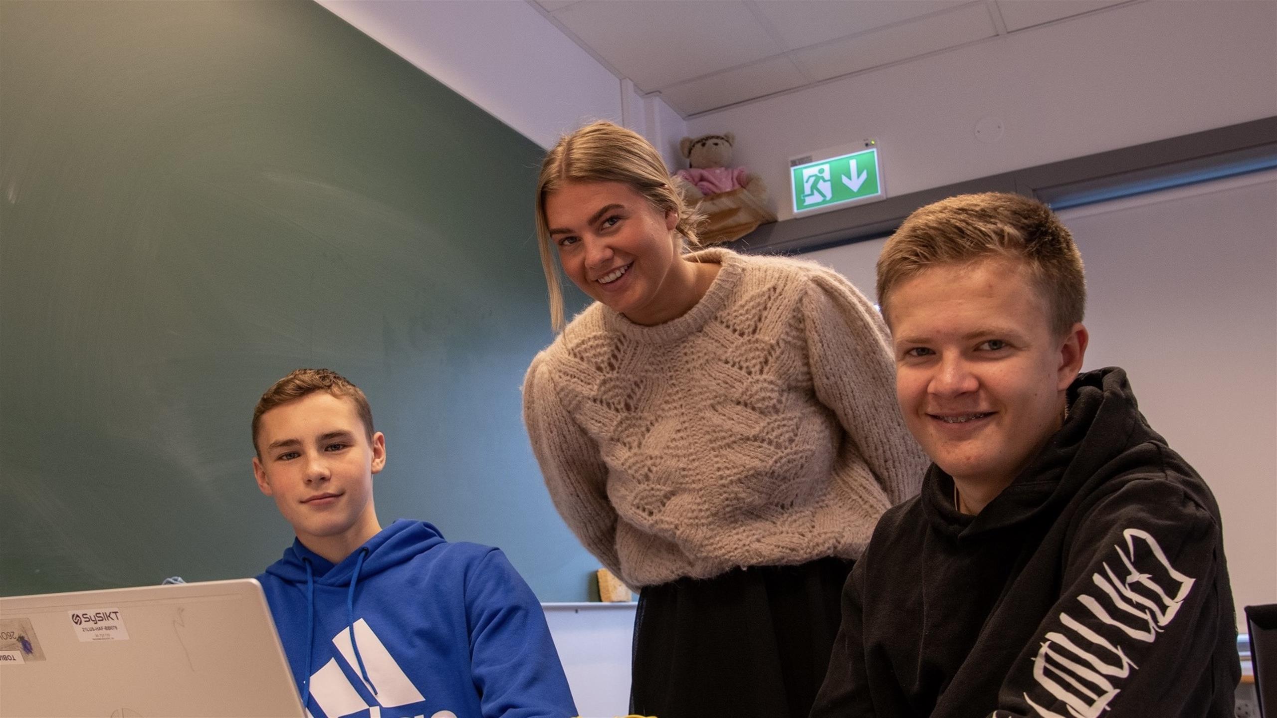 Tre smilende studenter avbildet i et klasserom.