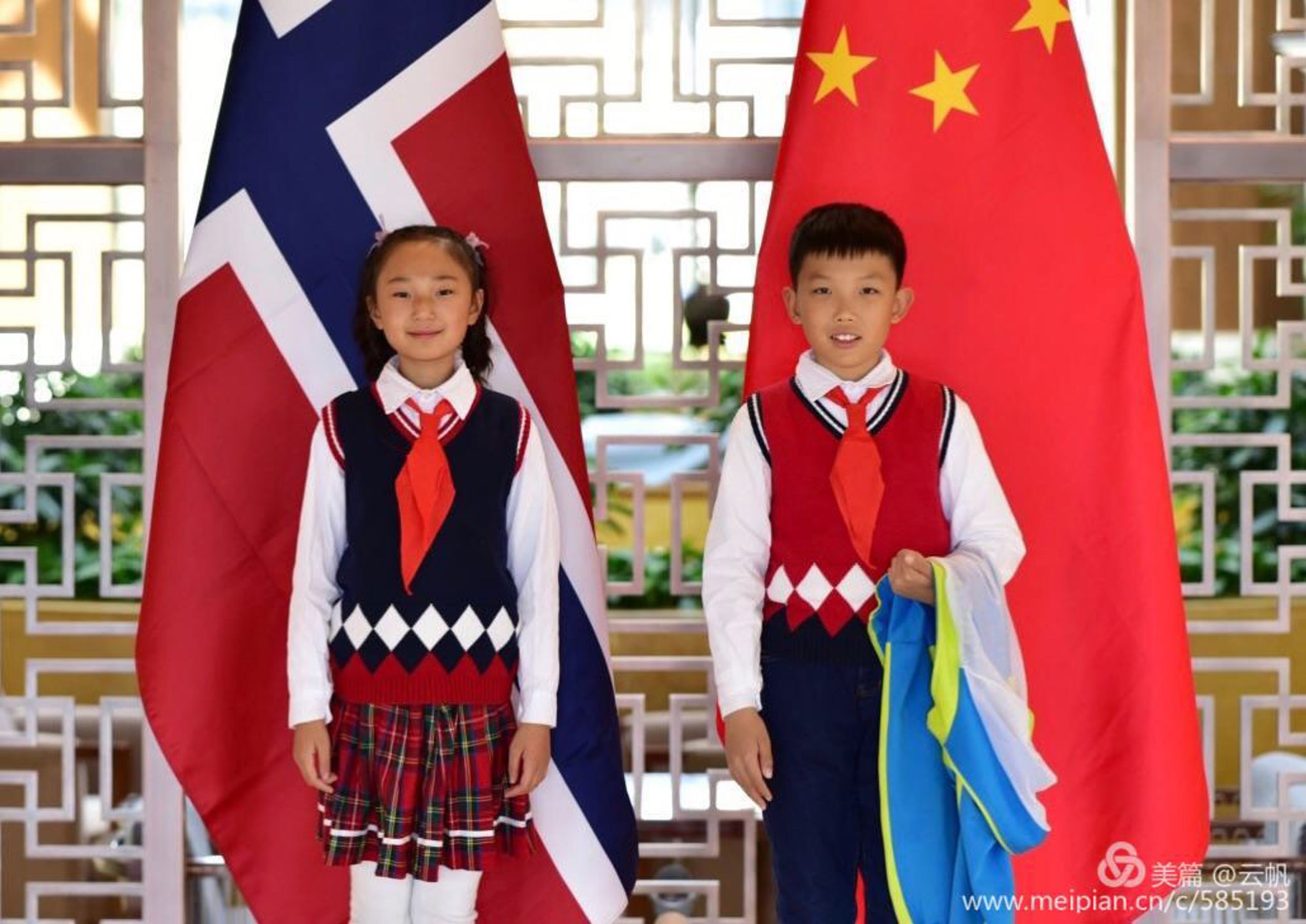 To children standing in front of a Norwegian and a Chinese flag. The child to the left is wearing a white shirt, a blue vest and a plaid skirt. The child to the right is wearing a white shirt, a blue pair of pants and a red vest.