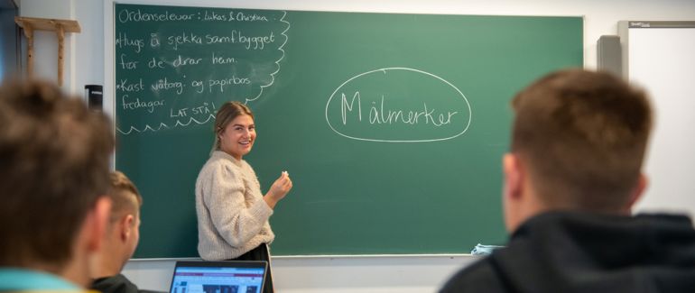 Lærer som står til venstre for tavlen foran en skoleklasse med kritt i hånden.