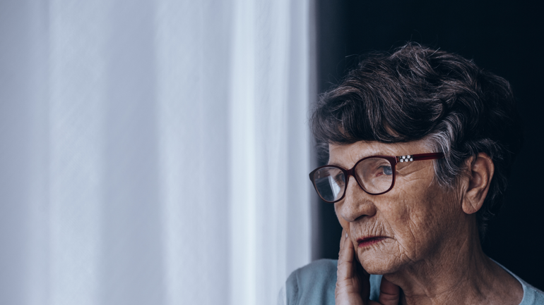 Eldre dame med briller står ved et vindu.