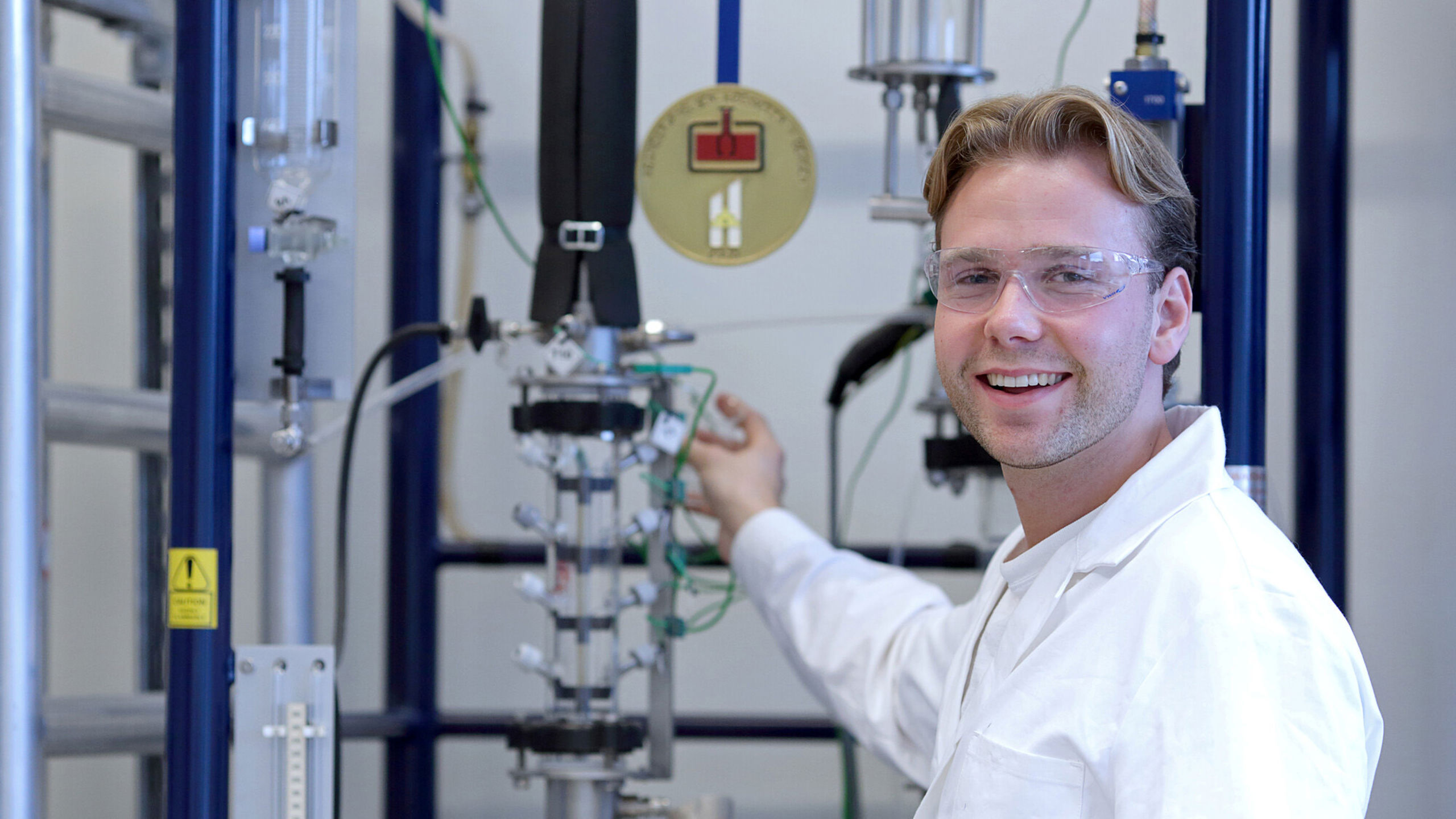 Ein blond person med kvit labfrakk står og smiler framfor noko teknologisk utstyr.