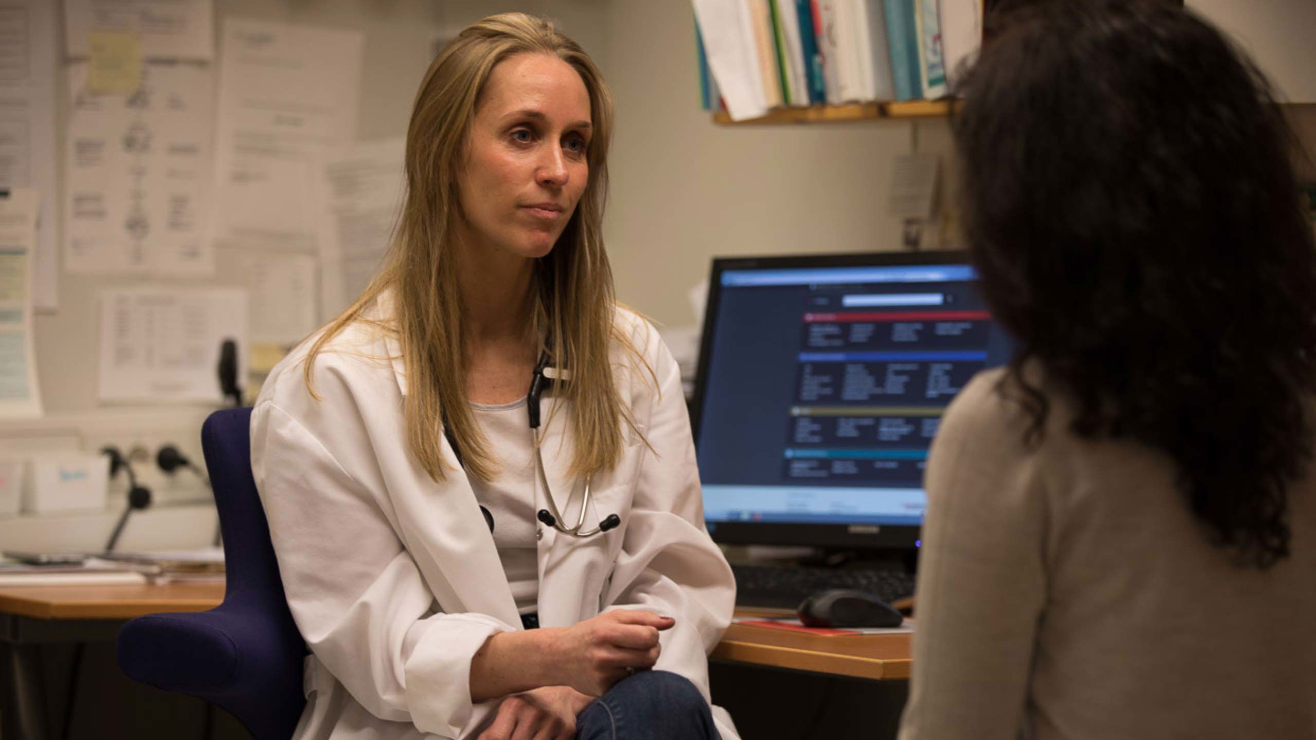 kvinne i helsefrakk sitter ved PC og snakker med pasient