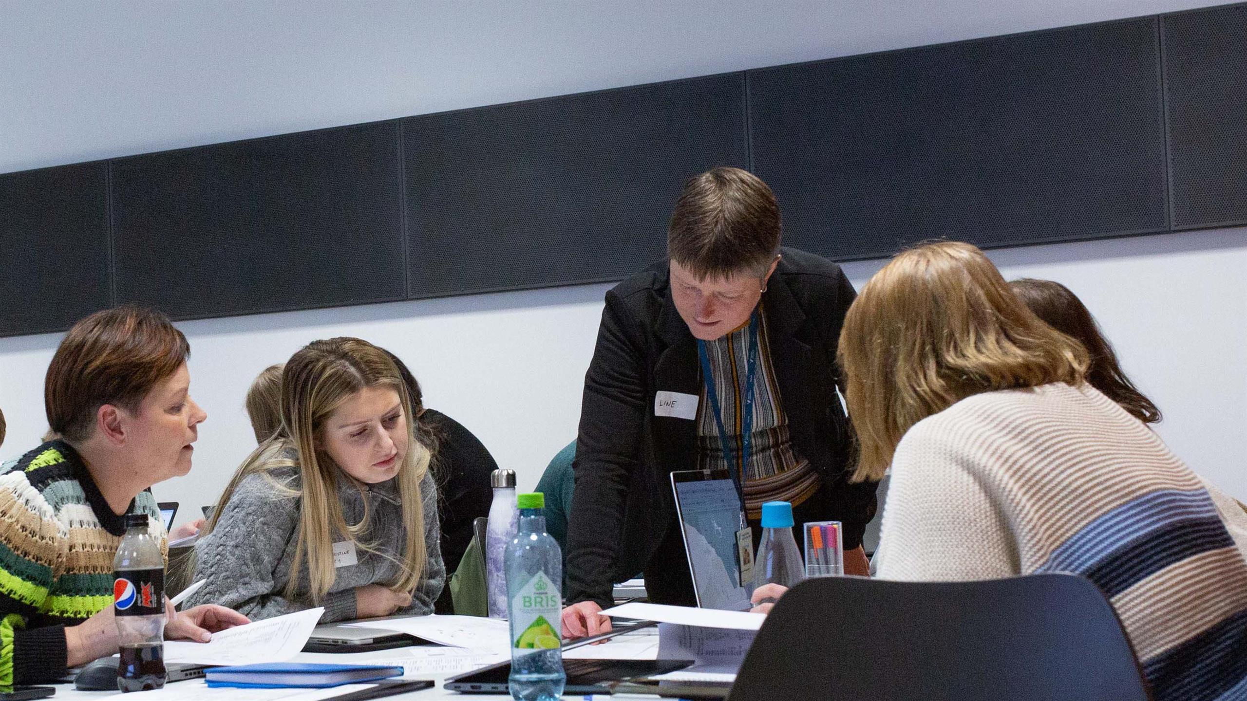 Gruppe mennesker jobber sammen rundt et arbeidsbord. En dame står over de og instruerer. 