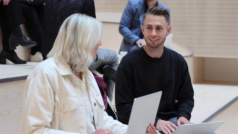 To personer sitter med bærbar pc på fanget. Mann ser inn i kameraet og smiler. 