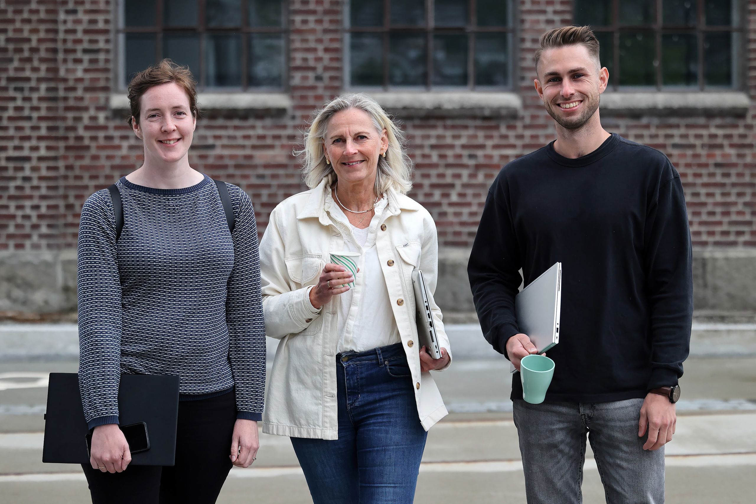 Tre voksne mennesker i ulik alder står utenfor en bygning. De har pc og kaffekopp i hånden.