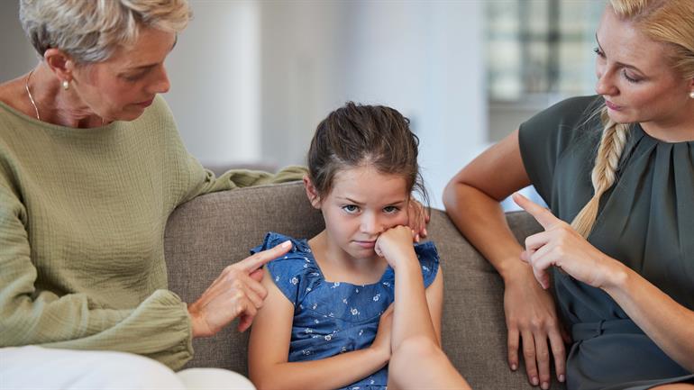 Jente sitter i sofa mellom to damer. Jenten ser nokså misfornøyd ut. 