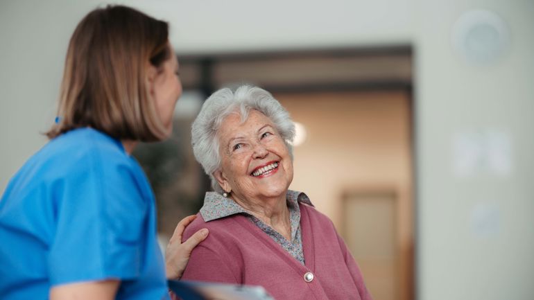 Eldre dame smiler og ser opp på en sykepleier.