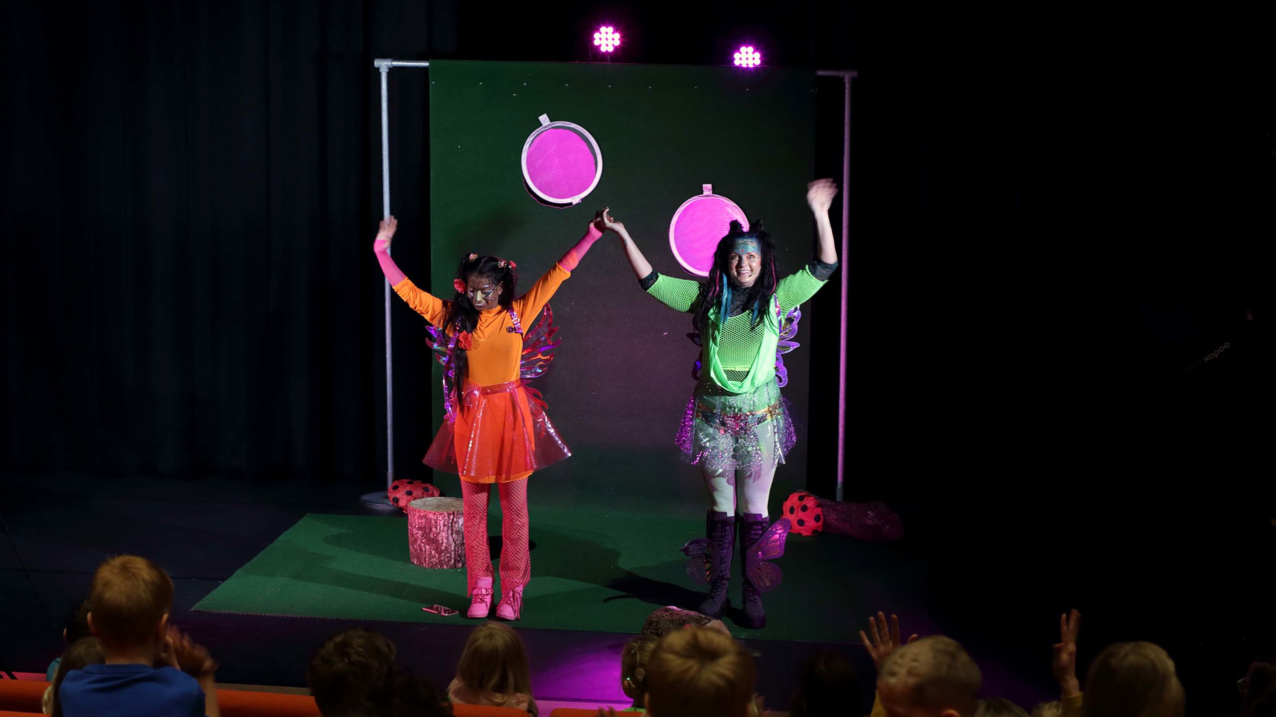 To skuespillere står på scenen foran barn. De har kostymer i sterke farger.