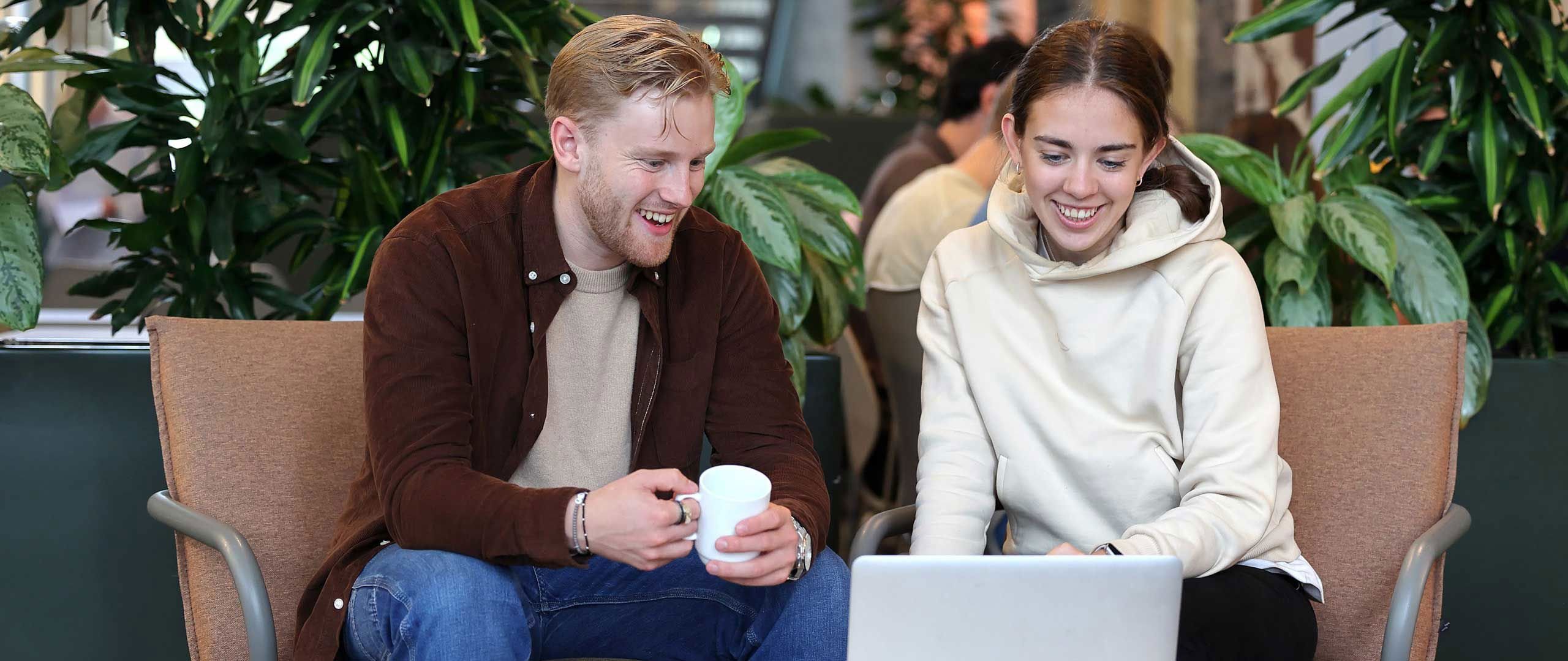 To smilande studentar som drikk kaffe og ser saman på ein datamaskin.