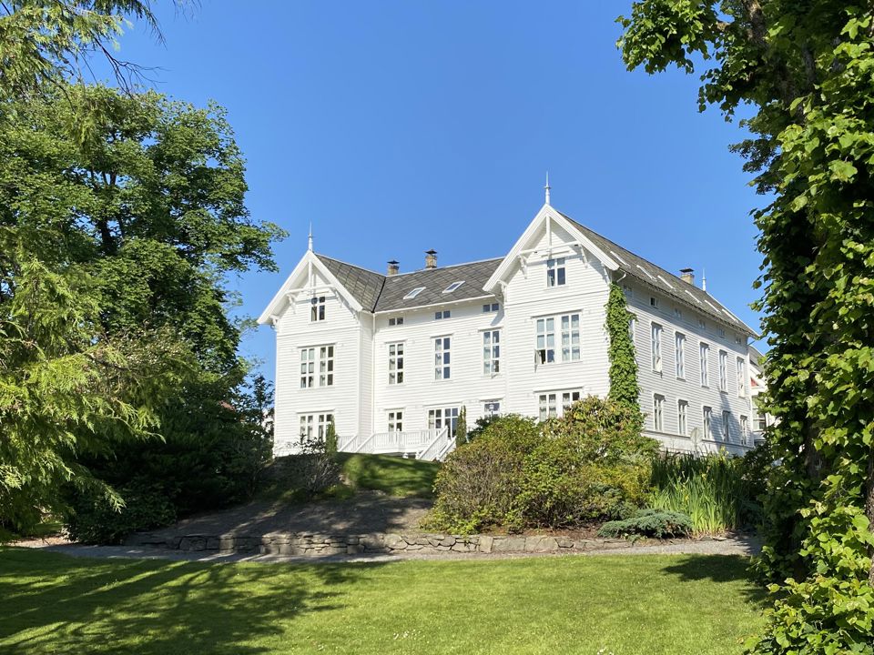 Bilde av eit stort, gammalt hvitt trehus, i ein grøn og frodig hage.