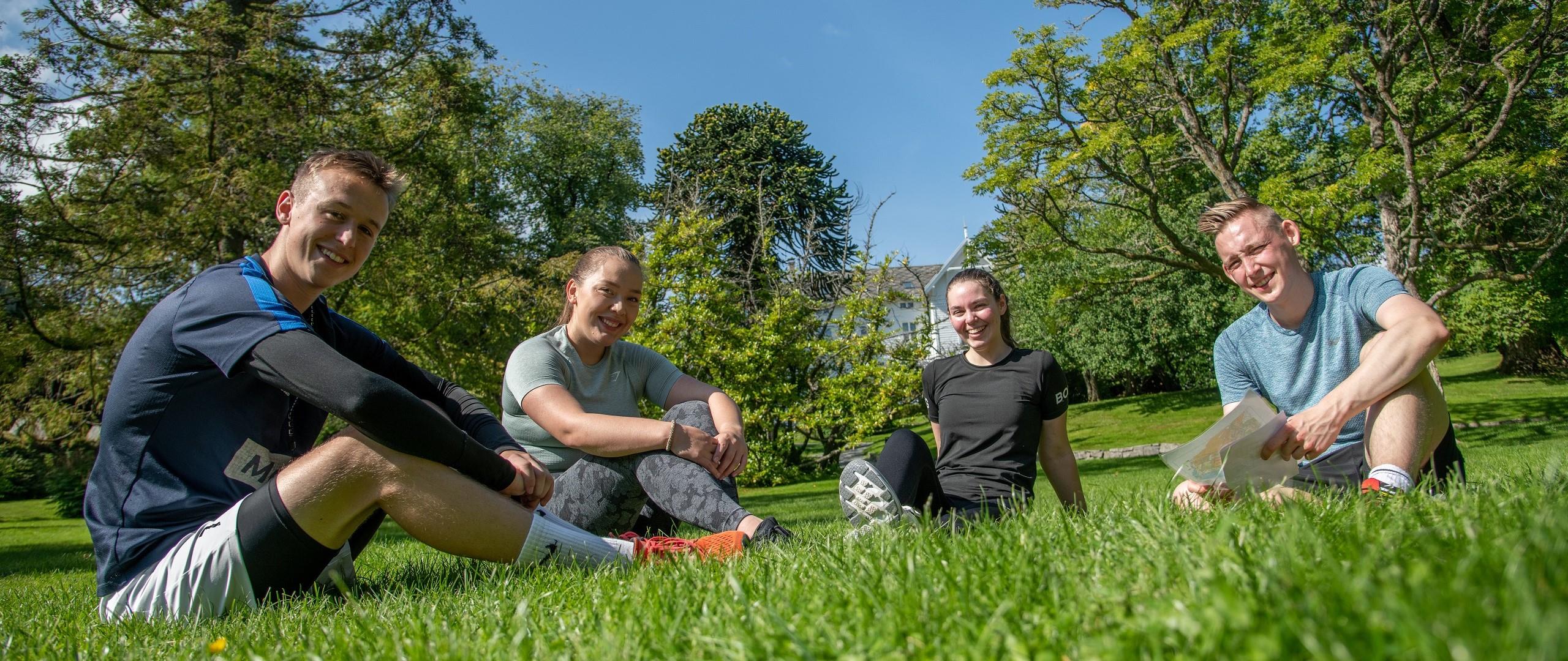 Fire studenter sitter i treningstøy på gresset og smiler til kamera.