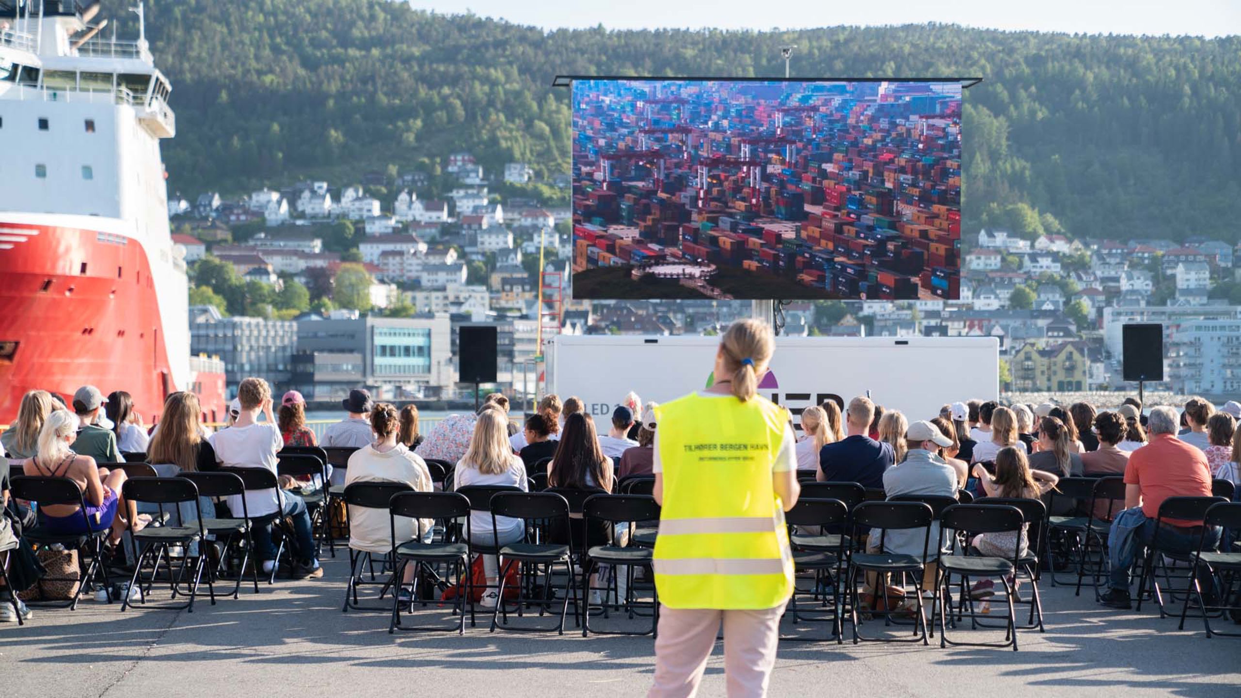Dame i gul vest står foran scene utendørs.