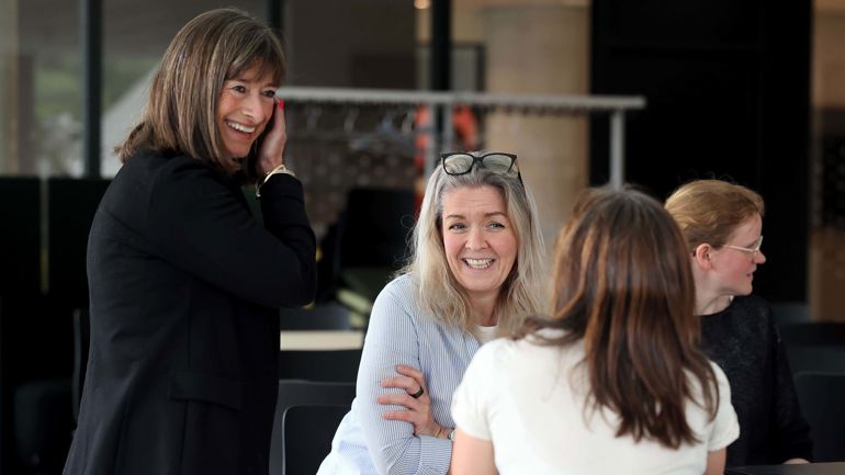 Gruppe sitter rundt bord. Kvinne smiler inn i kamera. En annen kvinne står bak og smiler.
