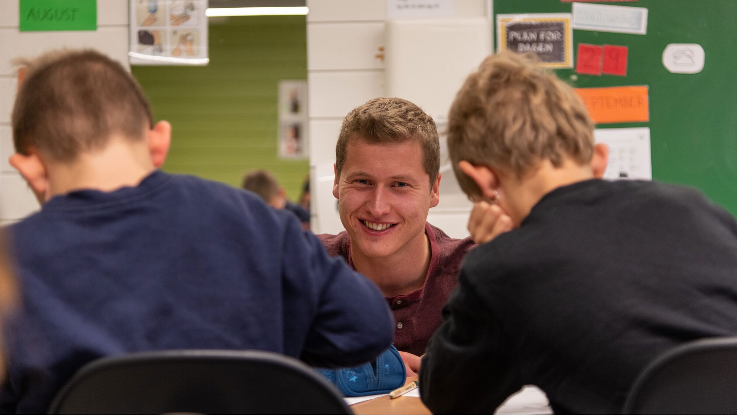 Mannlig lærer smiler mot kamera. Han sit i ein undervisningssituasjon med to elevar. Elevane sit med ryggen mot kamera, litt uklare på bildet