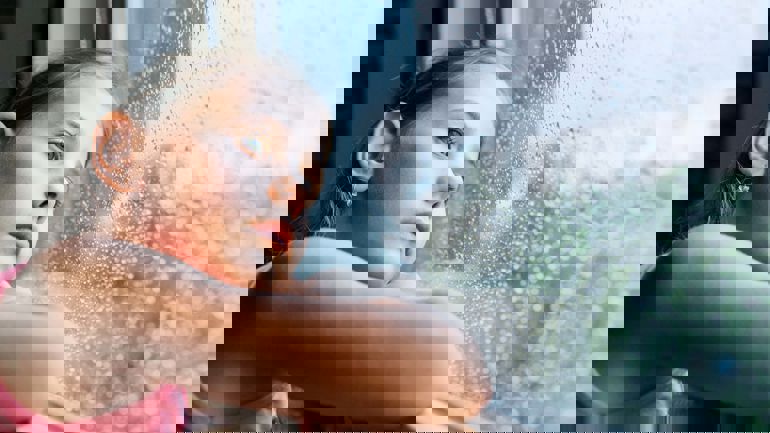 Jente sitter i vinduskarm og ser ut på regndråper på vinduet.