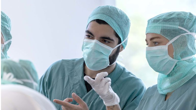 To sykepleiere med uniform, hårnett og munnbind ser på noe og peker.