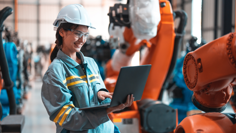 Kvinne med hjelm og vest står foran en stor maskin. I hendene har hun en pc.