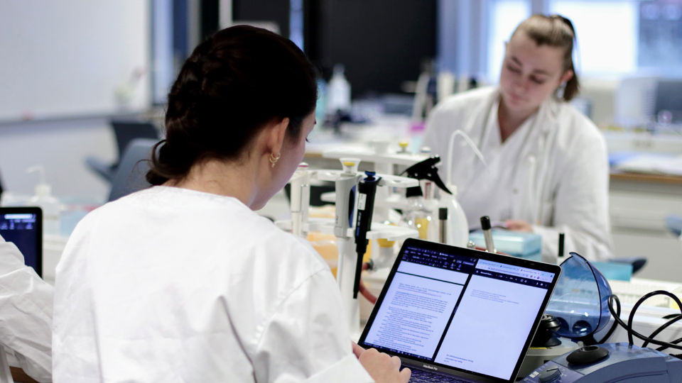 To studentar, kledd i kvitt, sit i ein lab. Den eine har ryggen til og ser på PC-en sin, den andre ser ned mot noko hen held i hendene, som vi ikkje kan sjå i bildet.