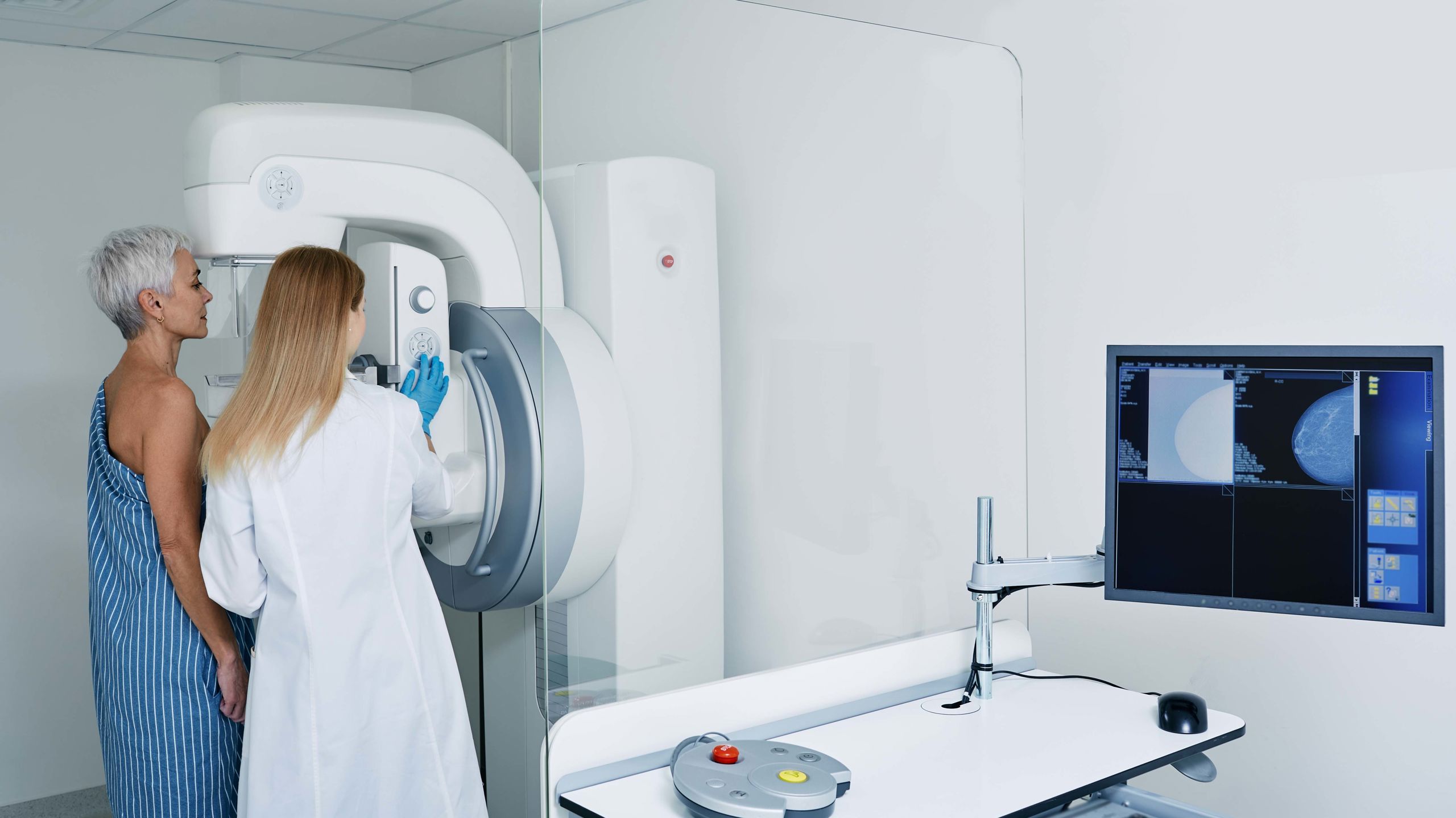 Dame i hvit frakk står sammen med halvt avkledd dame foran en stor maskin for å ta røntgen.