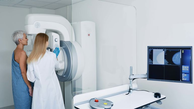 Dame i hvit frakk står sammen med halvt avkledd dame foran en stor maskin for å ta røntgen.