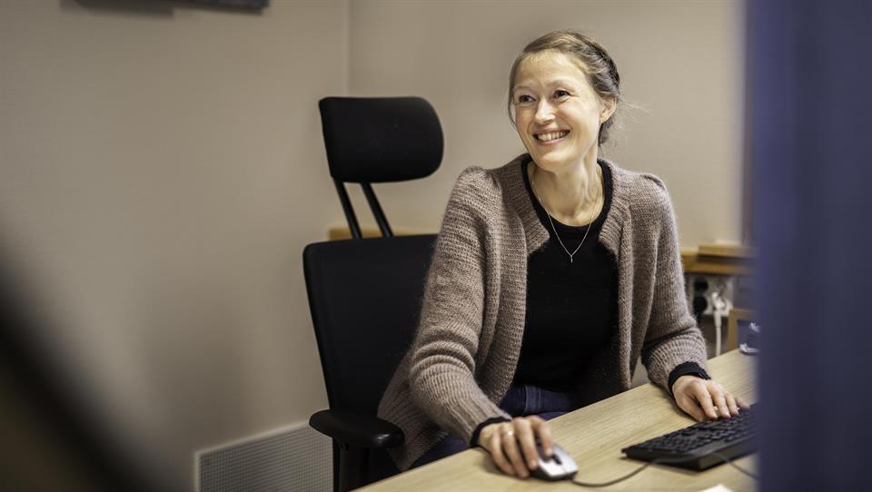 Kvinne sitter ved eit kontorbord framfor PC-en. Ho smiler og ser til sida, verkar å vere i samtale med ein person i venstre kan som vi ikkje kan sjå.