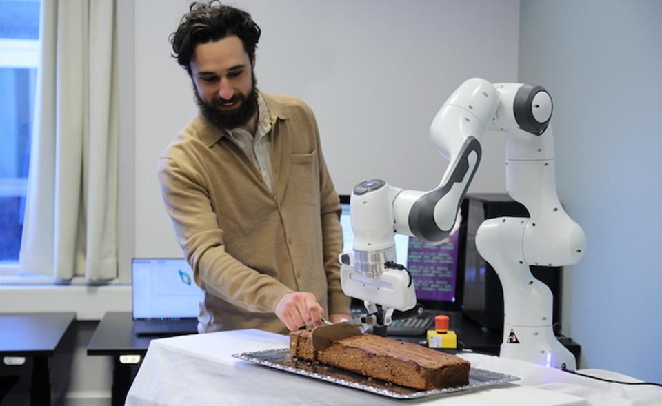 Daniel Schäle (stipendiat) står ved siden av en robotarm som skjærer opp en sjokoladekake på et bord.