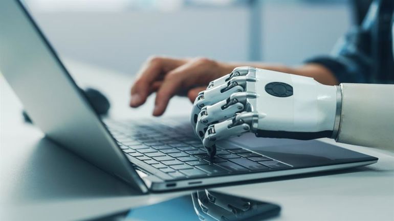 The image illustrates one human hand and one robotic hand in cooperation over the keyboard on a laptop computer.